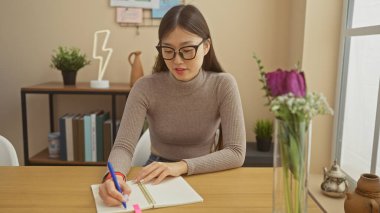 Genç Asyalı bir kadın rahat bir evde kalem ve defterle ders çalışıyor..