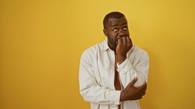 Yakışıklı, Afro-Amerikalı genç bir adam, parlak sarı bir arka plan karşısında, yalnız duruyor, beyaz gömlek giyiyor ve düşünceli bir ifade sergiliyor..
