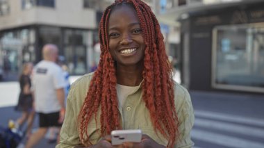 Gülümseyen Afro-Amerikan kadın. Kırmızı örgülü. Güneşli şehir caddesinde akıllı telefon kullanıyor.