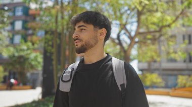 Sakallı genç bir adam güneşli bir şehir parkında düşünceli bir şekilde yürüyor, sıradan, şık bir tavır sergiliyor..