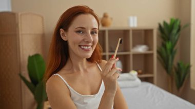 Young attractive redheaded woman in a spa wellness center holding a toothbrush while smiling in a serene indoor setting with plants and a calm beige interior background clipart