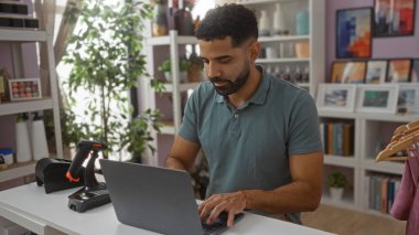 Ev dekorasyonu mağazasında dizüstü bilgisayarda çalışan genç bir adam. Etrafı çeşitli dekorasyonlar ve ev bitkileriyle çevrili..