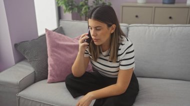 Genç yetişkin bir kadın, modern bir oturma odasında gri bir koltukta oturmuş endişeli bir ifadeyle akıllı bir telefonla konuşuyor..