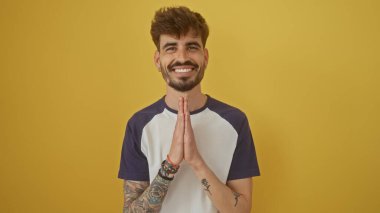 Dövmeli ve sakallı neşeli bir genç adam. Parlak sarı bir arka plana karşı el ele poz veriyor.