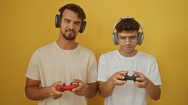 İspanyol baba ve oğul kulaklık takıyor, video oyunları oynuyor, sarı bir duvara karşı yan yana duruyor, bir anlık sevgi ve bağ paylaşıyorlar.