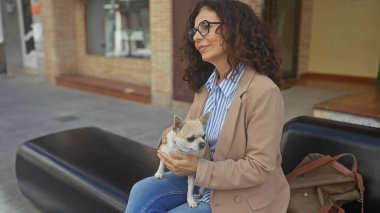 Orta yaşlı, İspanyol bir kadın, elinde köpeğiyle şehirde bir bankta oturuyor..