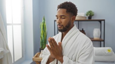 Sağlıklı bir kaplıcada meditasyon yapan genç bir adam, beyaz bir bornoz giyer, rahat ve sükunet saçar. İç mekan güzellik merkezinde sakin bir dekor ve doğal ışık sağlar.