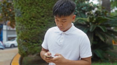 Şehir parkında beyaz polo giyen genç Çinli bir adam, arka planda yeşil ağaçlar ve binalar olan şehir dışındaki akıllı telefonuna bakıyor..