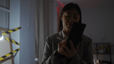 Asyalı bir kadın kapalı alanda loş bir suç mahallini araştırıyor, tabletine notlar alıyor..