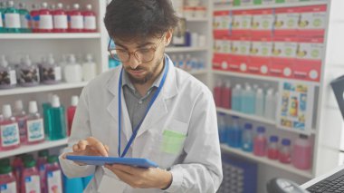 A young bearded man in glasses and a white lab coat focuses on a tablet inside a brightly lit pharmacy. clipart