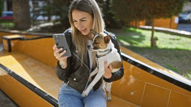 Genç bir kadın köpeğiyle dışarıda vakit geçirmekten hoşlanıyor, güneşli bir bankta selfie çekiyor..