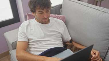 Genç bir İspanyol adam evinde kanepede rahat bir şekilde kucağında kedi uyuyan bir dizüstü bilgisayarla ev ortamını tasvir ediyor..