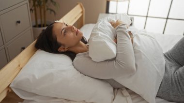 Genç, rahat bir yatak odasındaki yatakta uzanan bir kadın, düşünceli ve rahat görünürken yastığa sarılıyor..