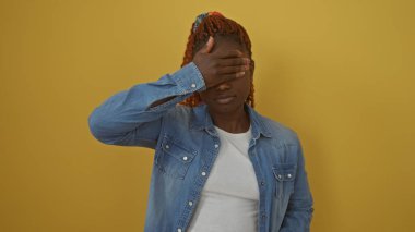 Kot ceketli Afro-Amerikalı genç bir kadın elini sarı bir arka plana koyup yüzünü kapatıyor.