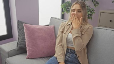 Genç, sarışın, beyaz bir kadın modern bir oturma odasında rahat bir kanepe ve dekoratif yastıklarla esniyor..