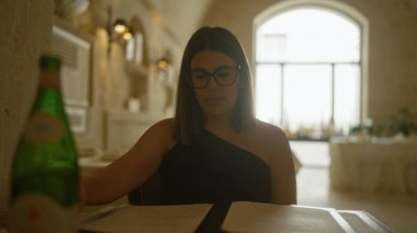 Kapalı bir İtalyan restoranında menüye bakan, loş ışıklandırmalı ve yanında bir şişe su olan bir masada oturan Latin kökenli genç bir kadın..