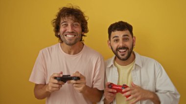Birlikte video oyunları oynayan iki adam, gülümseyen ve eğlenen, izole edilmiş sarı bir arka plana karşı, paylaşılan aktiviteler ve aile bağlarıyla dostluk ve bağlantı gösteriyorlar.