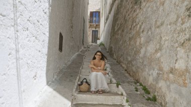 Güzel, genç bir İspanyol kadın, tarihi mimariyle çevrili eski bir kasaba olan Ostuni, Puglia, İtalya 'da basamaklarda oturur, sakin ve resimli bir Avrupa manzarası yakalar..