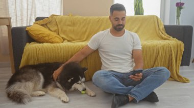 Genç İspanyol bir adam, rahat, aydınlık bir oturma odasında akıllı telefon kullanırken bir Husky 'yi okşuyor..