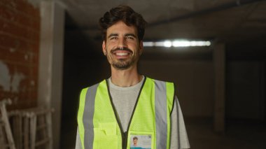 İnşaat alanında sakallı ve güvenlik yeleği olan gülümseyen genç bir İspanyol.