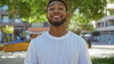 Yakışıklı bir adam yeşil ve şehir binalarıyla çevrili bir şehir parkında güvenle gülümsüyor. Açık hava hayatının özünü yakalıyor..