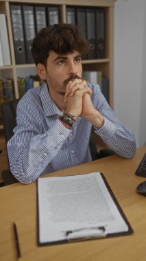 Ofiste sakallı, yakışıklı, genç bir adam. Not panosu ve bilgisayarın önünde düşünceli bir ifadeyle oturuyor.