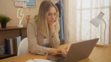 A young blonde woman works on her laptop in a cozy home office setting, surrounded by books and a modern lamp in a light-filled room. clipart