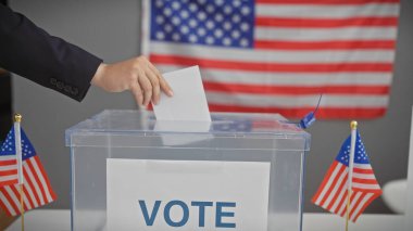 Amerika Birleşik Devletleri oy verme merkezinde, arka planda Amerikan bayrakları olan saydam bir sandıkta oy kullanan bir kişi..