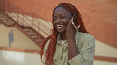 Genç bir Afro-Amerikan kadını güneşli bir sokak ortamında akıllı bir telefonla konuşmaktan hoşlanıyor..