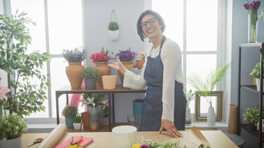 Parlak, kapalı bir çiçekçi dükkanında çeşitli bitkiler sunan gülümseyen kadın..