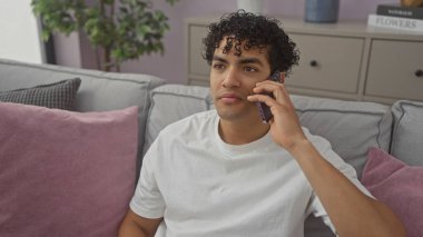 Genç bir İspanyol adam iyi dekore edilmiş bir oturma odasındaki koltukta otururken cep telefonuyla konuşuyor..