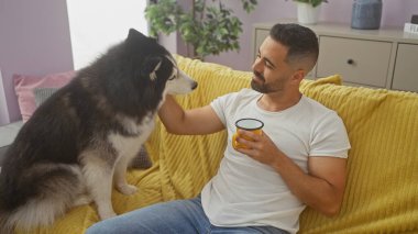 Genç bir İspanyol adam evde gevşiyor, sarı bir kanepede köpeğiyle bağ kuruyor, elinde kahve fincanı tutuyor..