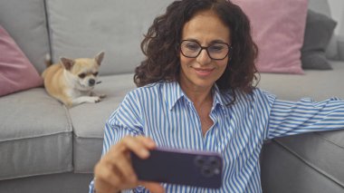 İspanyol kadın, oturma odasında Chihuahua 'nın yanında akıllı telefonuyla selfie çekiyor.