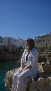 Genç ve güzel bir İspanyol kadın polignano 'da denize bakan kayalıklarda oturur kısrak, puglia, İtalya, geleneksel mimarisi ve arka planda açık mavi gökyüzü vardır..