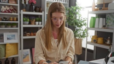 Arka planda çeşitli dekoratif eşya rafları olan bir ev dekoratörü dükkanında telefon karıştıran genç bir kadın.