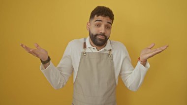 Confused young hispanic man with beard in apron shrugs shoulders against a yellow background clipart