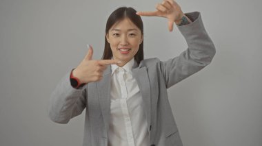 Gülümseyen Asyalı bir kadın ellerini beyaz bir arkaplana dayayıp yüzünü çerçeveliyor, takım elbise giymiş..