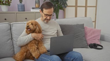 Latin kökenli orta yaşlı bir adam rahat bir oturma odasında dizüstü bilgisayar kullanırken bir kanişi kucaklıyor..
