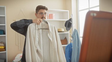 Genç bir İspanyol adam, iyi aydınlatılmış bir odada aynalı bir gömleği inceliyor, günlük moda seçimi senaryosunu tasvir ediyor..