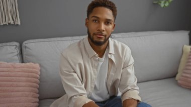 Modern bir oturma odasında rahat bir koltukta oturan yakışıklı genç adam. Tarafsız dekorları ve rahat yastıkları var.