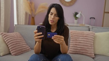 Modern dekorlu rahat oturma odasında akıllı telefon ve kredi kartı kullanan İspanyol kadın..