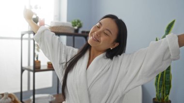 Çinli bir kadın spa salonunda dinlenmekten zevk alıyor, beyaz bir bornoz giyiyor, gülümsüyor ve kollarını kapalı bir sağlık merkezinde açıyor..