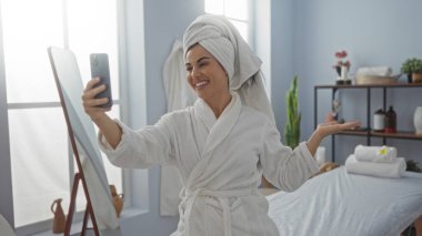 Genç bir kadın sağlık merkezinde video konferans veriyor, havlu ve bornoz giyiyor, spa ziyareti sırasında gülümsüyor ve sevinçle el sallıyor.