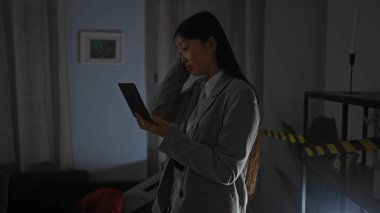 Oturma odasında genç bir Asyalı kadın, yoğun bir şekilde evde loş bir suç mahallinde tablet okuyor..