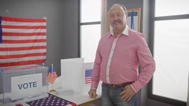 Bıyıklı orta yaşlı bir adam, Amerikan seçim sürecini sembolize eden ABD bayrağı ve oy sandığıyla bir odada duruyor..