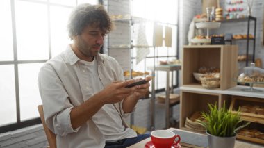 Genç adam fırının içinde akıllı telefon kullanıyor. Etrafı ekmek ve hamur işleriyle çevrili..