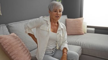 Evdeki bir kanepede oturan kısa gri saçlı ve sırt ağrısı çeken bir kadın.