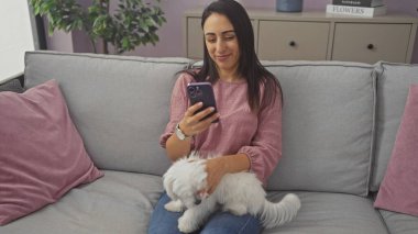 Beyaz köpeğiyle evde gri bir kanepede vakit geçiren İspanyol kadın akıllı telefonuna bakıyor..