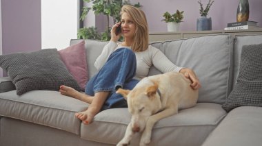 Rahat genç bir kadın, rahat bir oturma odasındaki kanepede uyuyan bir köpeği okşarken telefonda konuşur..