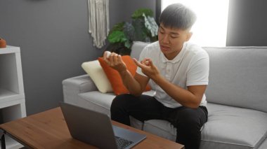 Genç adam modern bir oturma odasında bir kanepede oturuyor, reçeteli bir şişeyi inceliyor ve arka planda yastıklar ve ev bitkileri olan ahşap bir masada dizüstü bilgisayar kullanıyor.